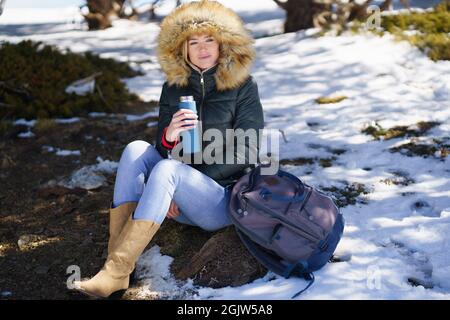 Femme buvant quelque chose de chaud d'une bouteille de thermos en métal assis sur un rocher dans les montagnes enneigées. Banque D'Images