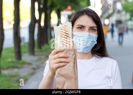 Une livreur masquée tient une commande - restauration rapide, nourriture de rue et livraison à domicile par messagerie pendant une pandémie. Banque D'Images