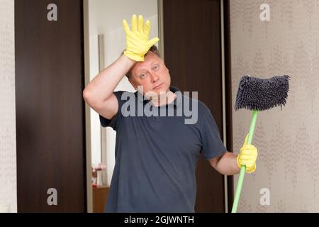 homme en gants de caoutchouc jaune avec une vadrouille dans ses mains frotte son front de la fatigue après le nettoyage de l'appartement. Banque D'Images