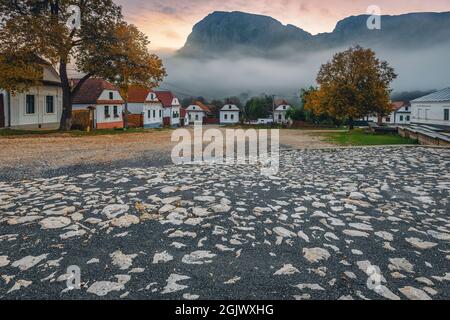 Superbe vue sur la rue rurale avec maisons traditionnelles blanchies à la chaux dans la rangée à l'aube. Village de Torocko et montagne Piatra Secuiului en arrière-plan au lever du soleil, Banque D'Images