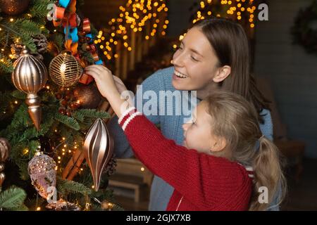 Bonne jeune mère aidant petite fille à décorer arbre de Noël. Banque D'Images