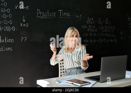 Souriant enseignant heureux ayant la leçon de mathématiques en ligne assis au tableau noir. Banque D'Images