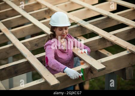 La jeune femme dans un casque de protection traite les planches sur le toit à l'aide d'une brosse Banque D'Images