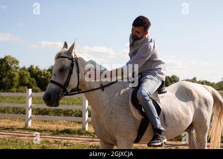 Le beau barbu monte un cheval blanc sur le ranch. L'homme fait le tour du cou du cheval Banque D'Images