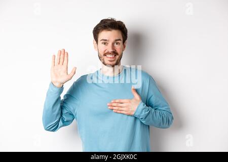 Homme souriant qui se soulève le bras et tient la main sur le coeur, faisant la promesse dire la vérité, jurer ou prêter serment, debout sur fond blanc Banque D'Images