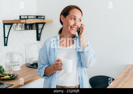 Jeune femme souriante attrayante avec une tasse de café dans la cuisine parlant au téléphone. Banque D'Images