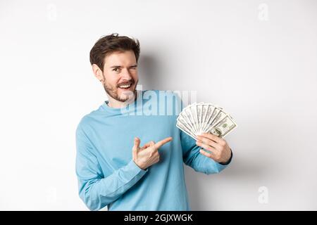 Beau gars riche qui se vante à la caméra, montrant des revenus, pointant le doigt sur l'argent gagné et souriant, debout sur fond blanc Banque D'Images