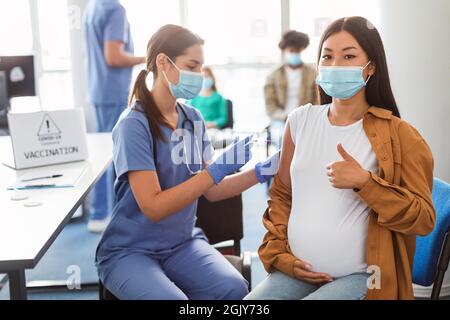 Femme enceinte au masque recevant le vaccin du coronavirus montrant des Thumbs Up Banque D'Images