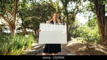 Femme à la mode regardant la caméra tout en tenant un écriteau vierge. Femme d'âge moyen affichant une bannière blanche. Militante féminine debout seule Banque D'Images