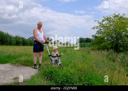 Vue en longueur d'un homme âgé avec une petite fille mignonne marchant dans la prairie verte. La petite fille est assise sur le vélo sur l'herbe verte Banque D'Images