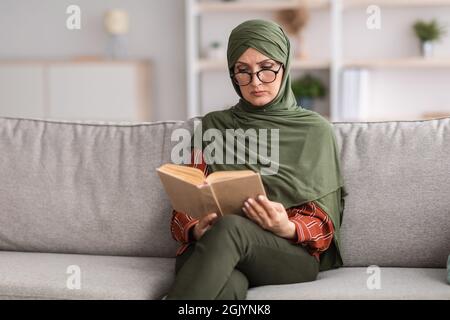 Livre de lecture de femme musulmane mature portant des lunettes assises à la maison Banque D'Images