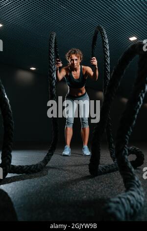 La jeune femme est engagée dans la salle de sport faisant des exercices crossfit avec des cordes de combat Banque D'Images