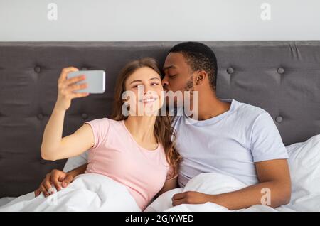 Jeune couple divers avec téléphone cellulaire embrassant tout en prenant le selfie au lit à la maison Banque D'Images
