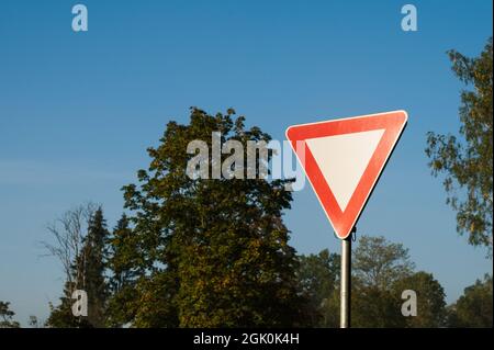 Gros plan du panneau de route de la voie de passage. Triangle rouge et blanc. Arbres verts et ciel bleu sur fond. Banque D'Images