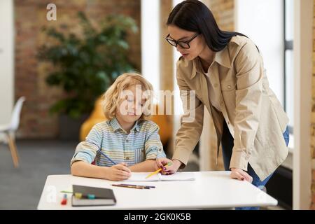 Jeune enseignante de soutien en lunettes aidant petit garçon avec la tâche. Enfant étudiant à l'école primaire, assis au bureau Banque D'Images