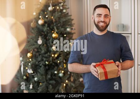 Portrait d'un jeune homme à barbe heureux dans un t-shirt debout avec cadeau contre l'arbre de Noël à la maison Banque D'Images
