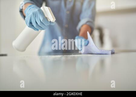 Gros plan des mains de femmes dans des gants en caoutchouc à l'aide d'un spray détergent et d'un chiffon tout en nettoyant la surface blanche de la cuisine Banque D'Images