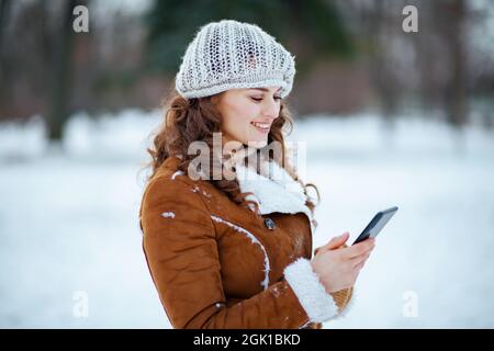 heureuse élégante femme de 40 ans à l'extérieur dans le parc de la ville en hiver dans un bonnet tricoté et un manteau de peau de mouton envoyant un message texte à l'aide d'un smartphone. Banque D'Images