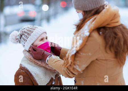 belle mère et fille dans un bonnet et manteaux tricotés en plein air dans la ville en hiver portant un masque médical. Banque D'Images