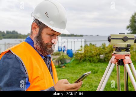 Un ingénieur civil avec un niveau optique regarde dans un téléphone portable. Un barbu recherche des informations sur son téléphone portable. Banque D'Images