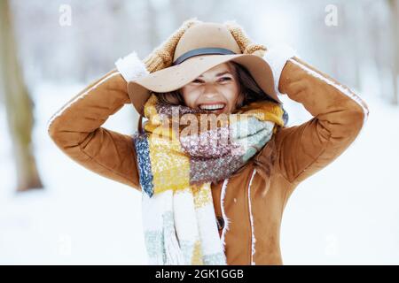 femme élégante souriante en chapeau marron et écharpe avec moufles en peau de mouton à l'extérieur dans le parc de la ville en hiver. Banque D'Images