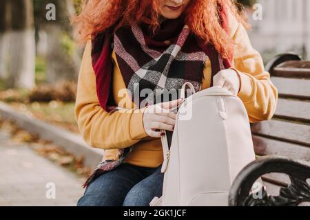 Jeune femme aux cheveux rouges en chandail jaune s'assoit sur le banc et cherche quelque chose dans son sac. Marchez le long d'une journée d'automne ensoleillée. Banque D'Images