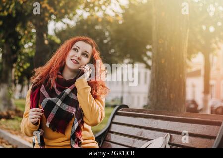 Jeune femme aux cheveux rouges en sweat-shirt jaune parlant au téléphone. Lady est assise sur le banc du parc le jour de l'automne. Style de vie. Banque D'Images