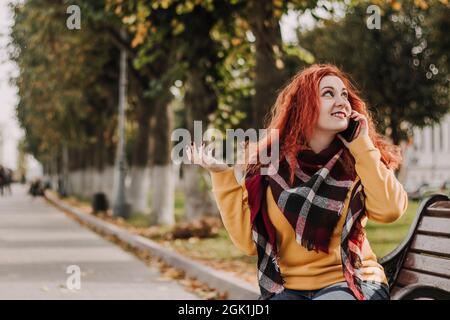 Jeune femme aux cheveux rouges en sweat-shirt jaune parlant au téléphone. Lady est assise sur le banc du parc le jour de l'automne. Style de vie. Banque D'Images