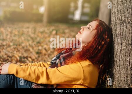 Femme aux cheveux rouges assise sur le sol, assise contre l'arbre et profitant d'une journée d'automne ensoleillée. Banque D'Images