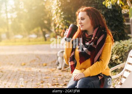 Une femme aux cheveux rouges en sweat-shirt jaune est assise sur le banc du parc et boit du café. Elle tient une tasse réutilisable dans ses mains. Portrait le jour d'automne ensoleillé. Li Banque D'Images