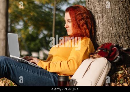 Jeune femme à cheveux rouges freelance travaille dans le parc à l'aide d'un ordinateur portable. Femme d'affaires moderne parlant au téléphone. Travail en ligne sur Internet. Banque D'Images