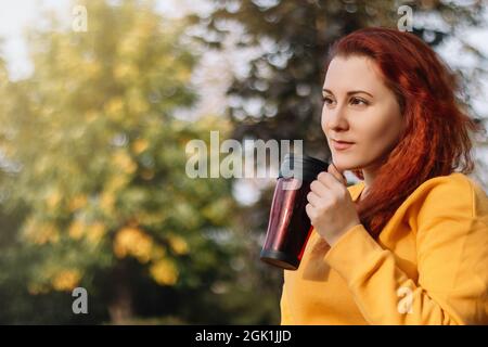 Une jeune femme aux cheveux rouges boit du café dans une tasse thermo réutilisable. Profitez de la journée ensoleillée d'automne dans la nature. Style de vie écologique. Banque D'Images