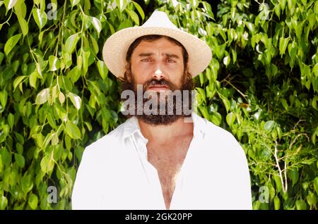 un jeune homme à la barbe et aux cheveux longs porte un chapeau et une chemise blanche entourée de verdure Banque D'Images