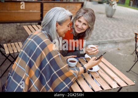 Une femme asiatique mûre montre son téléphone mobile à son meilleur ami dans un café de rue le jour de l'automne Banque D'Images