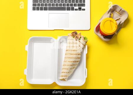 Boîte à lunch en plastique avec de savoureuses fajitas, un ordinateur portable et une tasse de café sur fond jaune Banque D'Images