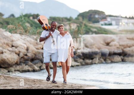 Bonne course mixte, couple d'âge moyen embrassant tout en marchant sur la plage tenant une guitare. Homme tenant guitare avec femme. Banque D'Images