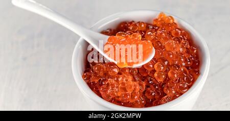 banderole avec un grand caviar rouge, délicieux et friable, dans une tasse blanche avec une cuillère sur fond blanc. Mise au point douce Banque D'Images