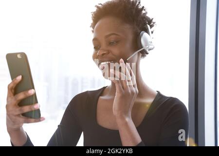 Gros plan d'une femme afro-américaine, parlant avec un casque sur une liaison vidéo. Une jeune femme sourit dans un uniforme sportif sur le fond de la fenêtre. Photo de haute qualité Banque D'Images