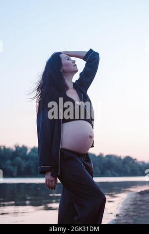 Brunette pose sur fond de nature. Belle jeune femme enceinte en pyjama de soie sombre pose et touche ses cheveux longs bruns soufflant dans Banque D'Images