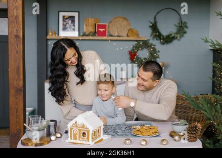 La famille se fait une maison de pain d'épice pour Noël avec des bonbons, le père met le glaçage sur le nez de son fils, ils rient Banque D'Images