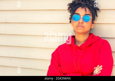 Fille adolescente cool adolescent mixte race biracial Afro-américaine femelle jeune femme portant des lunettes de soleil bleues et un sweat à capuche rouge vacances en été soleil Banque D'Images