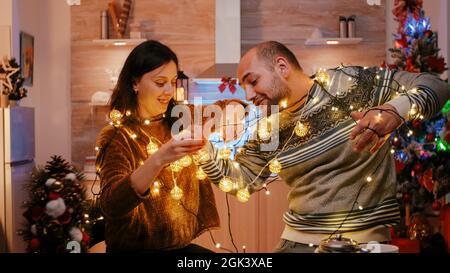 Couple joyeux s'emmêle dans une série de lumières scintillantes pour les préparatifs de la veille de noël. Homme et femme riant tout en démêlant nœud de guirlande avec des lumières et des ampoules illuminées Banque D'Images