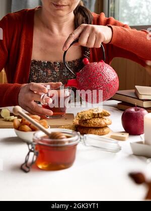 Une femme portant un chandail orange verse un thé chaud de la théière rouge dans une tasse en verre sur la table. Miel, biscuits et livres près de la tasse de thé sur le linge t Banque D'Images