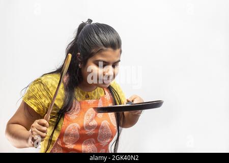 Une jolie femme de ménage indienne dans un tablier de cuisson avec une spatule en bois et une poêle à frire dans les mains sur fond blanc Banque D'Images