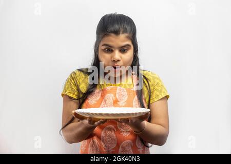 Une jolie femme de ménage indienne portant un tablier de cuisine avec une assiette de service à la main regarde dans la surprise sur fond blanc Banque D'Images