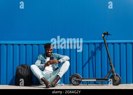 Beau Afro-américain assis sur un scooter électrique, avec un téléphone dans ses mains, et en tapant un SMS, 5g Internet, correspondance en ligne, ag Banque D'Images