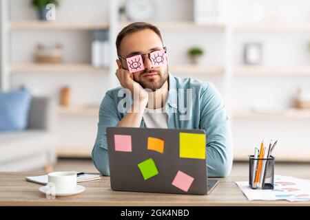 Jeune homme fatigué ayant des autocollants avec des yeux ouverts dessinant sur des lunettes, dormant près d'un ordinateur portable, prétendant travailler au bureau Banque D'Images