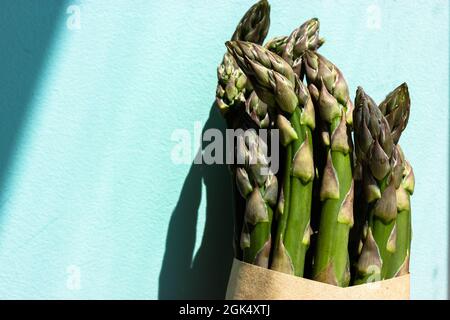 Un bouquet, un paquet de pousses d'asperges vertes fraîches enveloppées dans un papier kraft brun, un sac écologique sur fond bleu clair avec des nuances contrastées. Usefu Banque D'Images