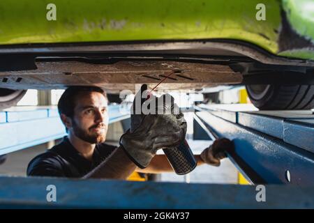 Gros plan d'un jeune homme, mécanicien automobile masculin dans des salopettes faisant le service de voiture à la gare, garage, intérieur. Concept du travail, de la profession, des affaires Banque D'Images