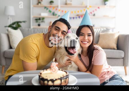 Joyeux couple multiracial célébrant l'anniversaire de leur chien avec un gâteau de fête, portant des chapeaux de fête à la maison Banque D'Images
